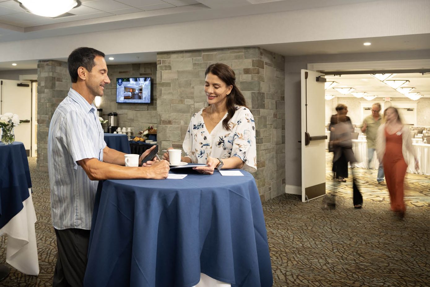 Two people chatting at a meeting table while other colleagues file into the meeting room